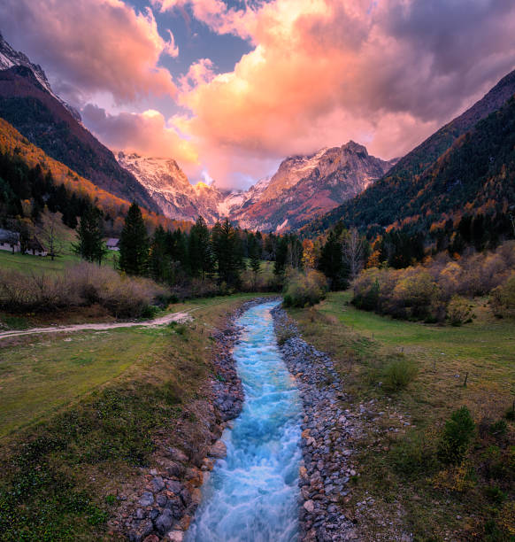 Aerial view of a blue mountain stream in green valley with autumn forests and snow-capped peaks at sunset. Top view of river with blue water, rocks in snow, trees and colorful cloudy sky in fall. Alps