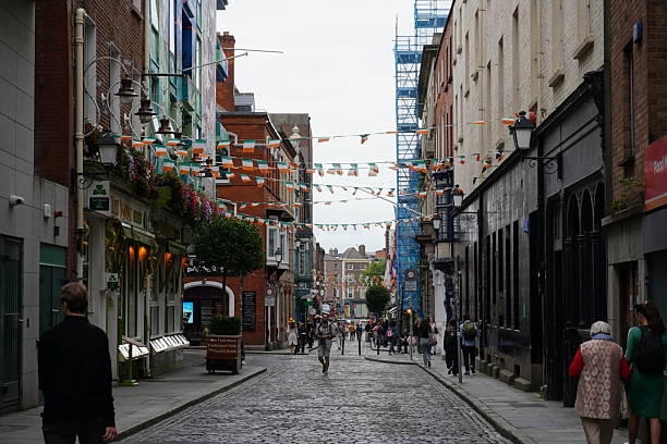 A narrow, cobblestone street in Dublin's city center is decorated with Irish flags and bunting, with people walking along the street.