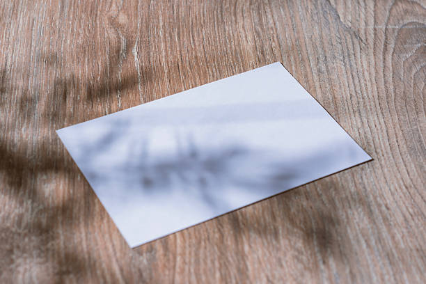 istock Minimalistic white mockup of a blank postcard lying on a wooden surface with soft natural shadows from plants. Template for branding, stationary design, wedding invitations and greeting cards. 2232924800