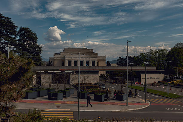 Geneva, Switzerland, Europe – April 9, 2025: General view of the Palais des Nations, the United Nations headquarters in Geneva, with the Alps in the background. The historic building is seen under dramatic spring clouds, while cars and pedestrians move through the entrance area in the foreground.
