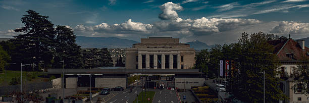 Geneva, Switzerland, Europe – April 9, 2025: General view of the Palais des Nations, the United Nations headquarters in Geneva, with the Alps in the background. The historic building is seen under dramatic spring clouds, while cars and pedestrians move through the entrance area in the foreground.