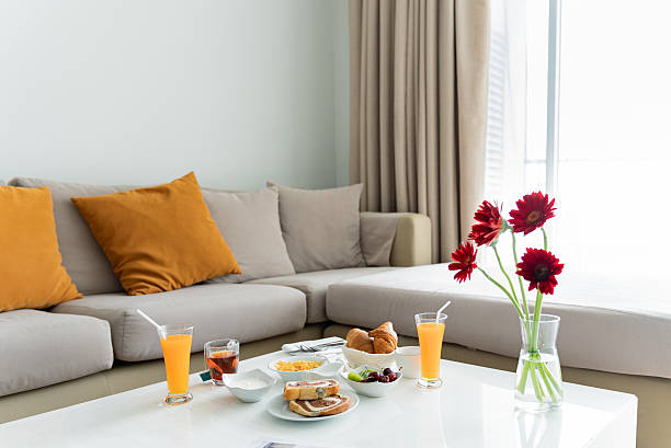 Upscale in-room dining breakfast arranged on a coffee table in a bright, modern hotel suite. The elegant spread features croissants, cereal, yogurt, fresh fruit, and drinks including orange juice and tea, with a newspaper and flowers adding a touch of comfort and relaxation. A sunlit sofa and neutral tones create a warm, inviting hotel atmosphere.