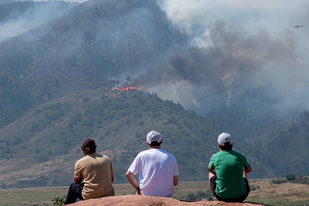 Jefferson County, Colorado (07/31/2024) -  A group of men sit on a rock watching the flames firefighting activities. A firefighting helicopter works to manage the Quarry Fire in Deer Creek Valley. The helicopter comes through the smoke and drops water from the air to manage the spread of the fire. As seen from South Valley Road in the Ken Caryl Ranch neighborhood on July 31 in Jefferson County.