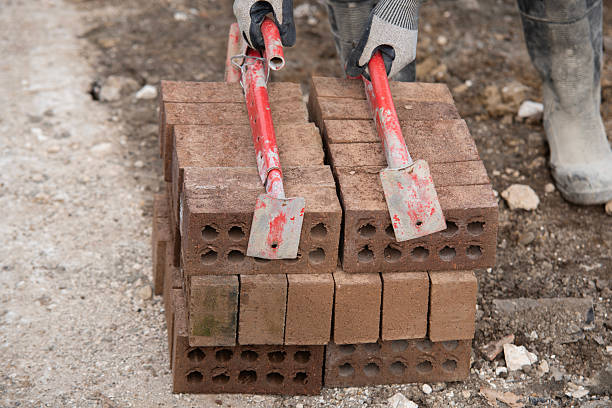 Bricklayer carrying red bricks on construction site with help of brick tongs lifting carrier