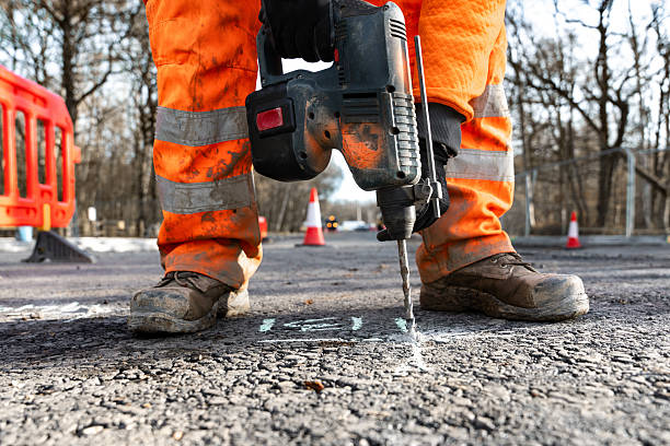 A local authority council workman in orange high visibility work clothes, using a breaker hammer to break up the tarmac on a city street. He is working on the edge of the street, where double yellow lines are painted, lifting big lumps of road material ahead of public road repairs.