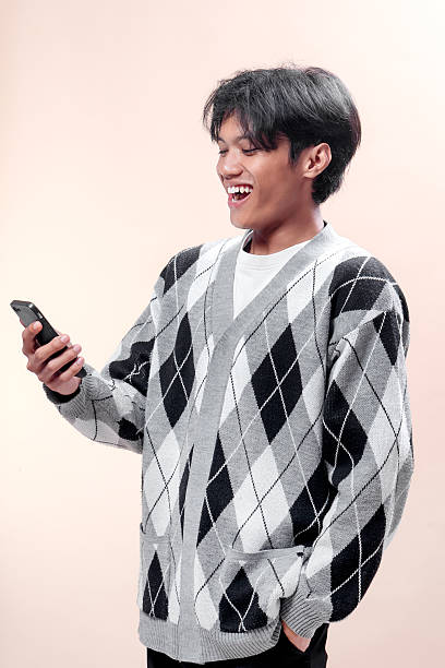 istock Asian man smiling while looking at smartphone, wearing casual argyle sweater, showing happiness and positive emotion, modern lifestyle and digital connection. 2232968637