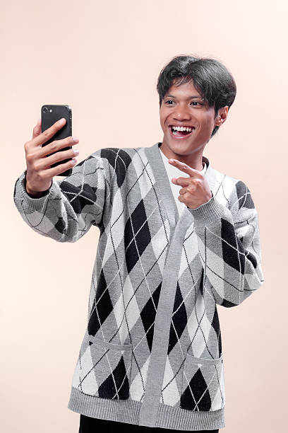 Asian man smiling brightly while holding smartphone and making finger gesture, showing cheerful energy, confidence, and modern lifestyle, perfect for advertising, business, and technology themes.