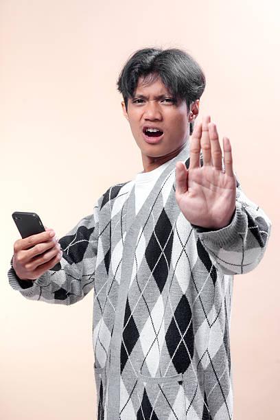 istock Asian man holding smartphone with surprised expression, raising hand forward as stop gesture, symbolizing rejection, warning, communication, modern lifestyle, and business concept. 2232968975