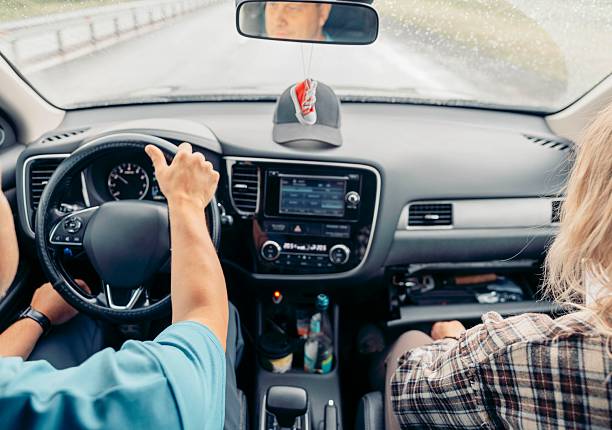 driver holding steering wheel inside modern car while passenger sits in front seat on rainy day road trip, interior dashboard, navigation screen, rearview mirror reflection, travel, transport