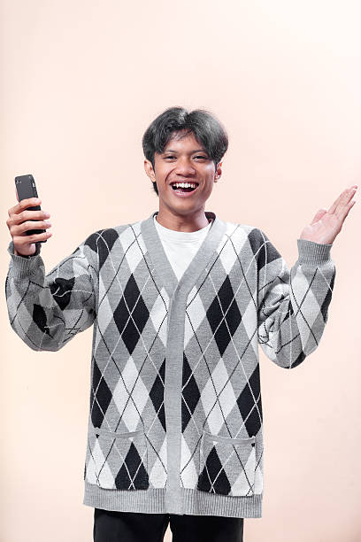 istock An Asian man smiling happily while holding a smartphone in one hand and gesturing with the other, wearing a gray argyle sweater, showing joy and confidence. 2232970128
