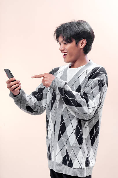 istock An Asian man smiling while pointing at his smartphone with excitement, wearing a gray argyle sweater, showing positive expression, modern lifestyle, technology use, and confidence. 2232970194