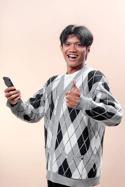 istock Asian man smiling confidently while holding a smartphone and showing a thumbs up gesture, wearing a gray argyle sweater, representing success, positivity, communication, and lifestyle. 2232970224
