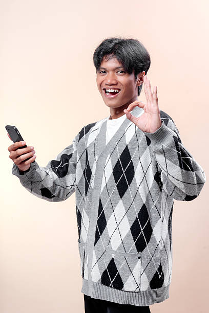 istock An Asian man smiling confidently while holding a smartphone and showing an OK hand gesture, wearing a stylish argyle sweater, expressing positivity and success. 2232970228