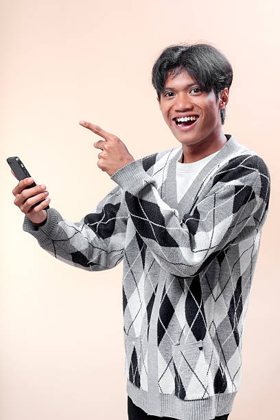 istock An Asian man smiling brightly while holding a smartphone and pointing his finger upward, wearing an argyle sweater, showing excitement and confidence. 2232970242