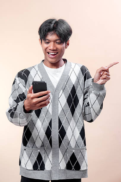istock An Asian man smiling while holding a smartphone and pointing sideways with his finger, wearing an argyle sweater, showing cheerful mood and confidence. 2232970278