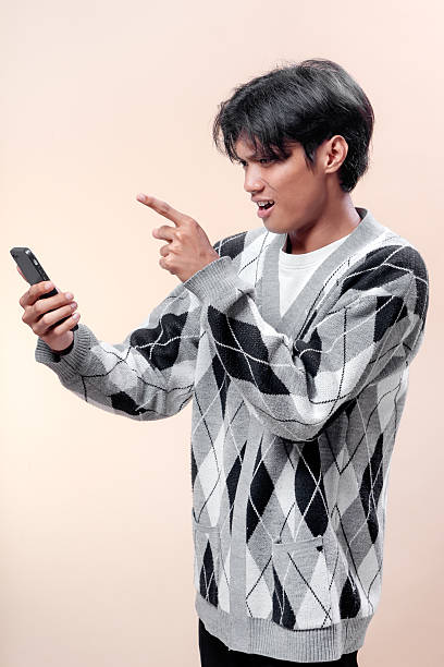 istock An Asian man looking at a smartphone with a surprised expression, pointing his finger at the screen, wearing a patterned sweater, showing curiosity. 2232970281