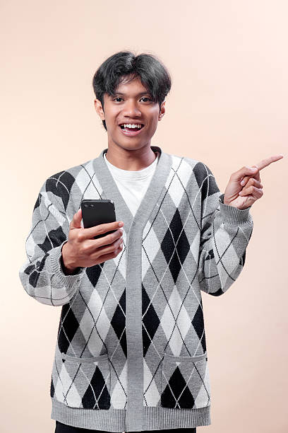istock An Asian man smiling while holding a smartphone and pointing his finger sideways, wearing an argyle sweater, showing cheerful expression and confidence. 2232970283