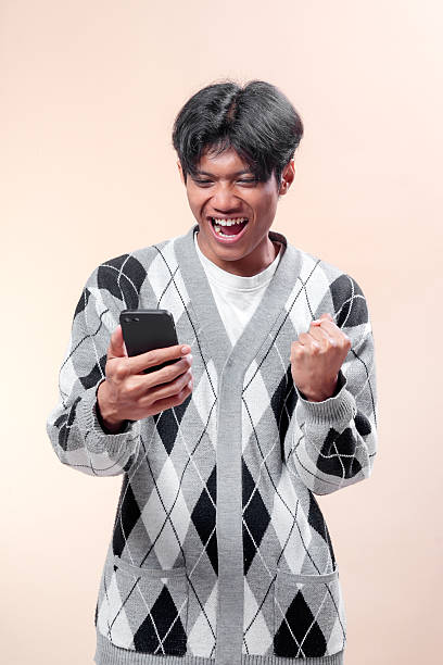 istock Asian man looking excited while holding smartphone, raising his fist with joyful expression, wearing gray argyle sweater, showing happiness, success, and positive energy. 2232970298