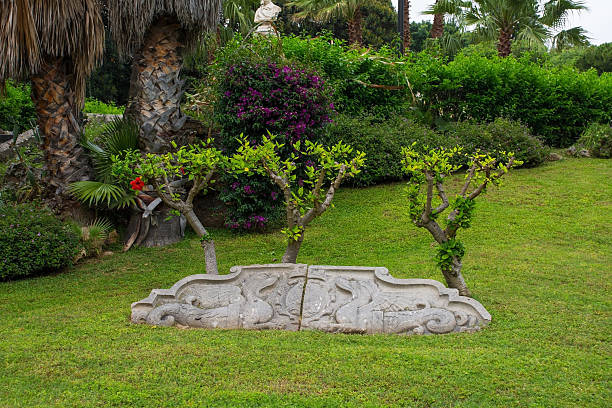 Piersanti Mattarella Park, originally called Giardino Inglese, in Liberta district, Palermo, Sicily, Italy. A large stone Baroque carving from an historic building, broken in two and depicting 2 stylized dragon-like sea creatures flanking a central shield or cartouche, now repurposed as a landscape decoration. Behind are 3 small pruned deciduous Hibiscus trees, one with a single red flower. In the background, Bougainvillea and palm trees