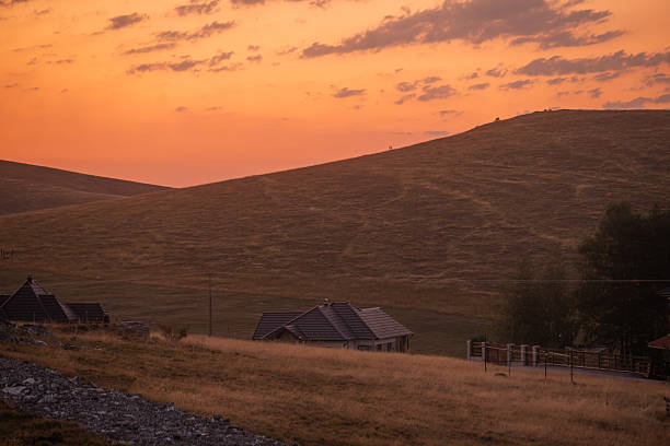Scenic view of a small village surrounded by rolling golden hills and dramatic mountain landscape at sunset.