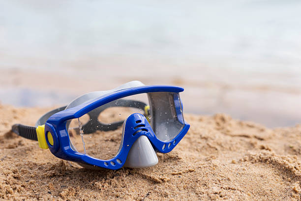A blue diving mask sits on warm sand at a beach during sunset. Gentle waves lap the shore, creating a peaceful ambiance perfect for water activities.