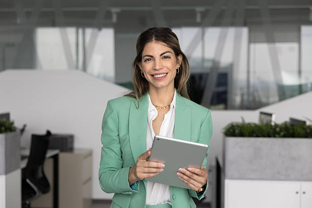 Beautiful Hispanic businesslady dressed in elegant jacket holds digital tablet posing alone in contemporary workspace, look at camera with joyful, positive expression. Modern innovative tech, workday