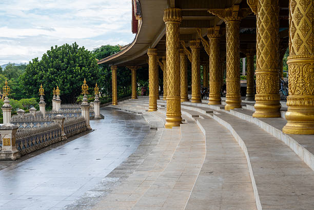 A majestic perspective shot showcasing a row of large, golden pillars with intricate, carved patterns. The columns support a curved roof, typical of traditional East or Southeast Asian architecture. The structure is situated on a clean, tiered stone walkway that leads to a decorative railing and lush greenery in the distance. The image conveys a sense of grandeur, cultural heritage, and peaceful beauty.
