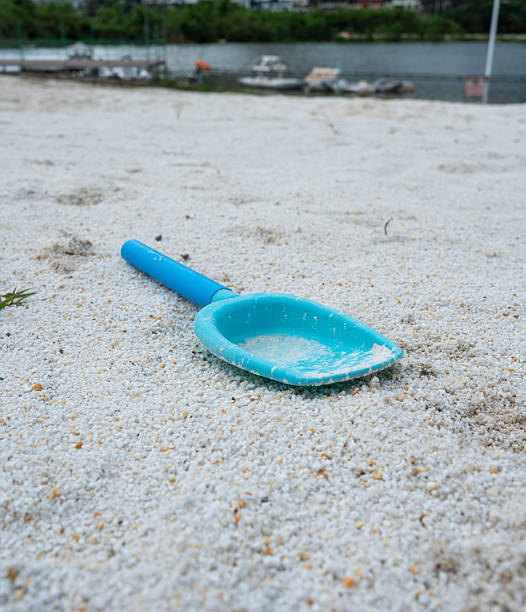 A simple, low-angle shot of a bright blue plastic shovel resting on a light-colored beach with granular sand. The foreground is in sharp focus, while the background is softly blurred, showing a body of water and distant land or boats. The image evokes a sense of nostalgia for childhood and summer vacations, suggesting a quiet, peaceful moment on a deserted beach after a day of play.
