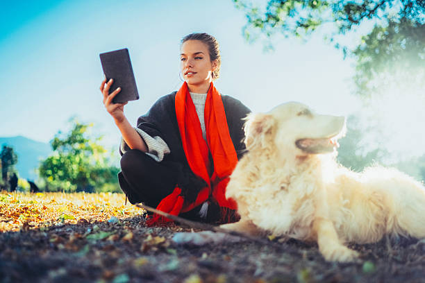 Woman enjoys reading from her electronic book while sitting on the ground next to her fluffy golden dog in a sunlit autumn park. The peaceful natural setting, vibrant scarf, and fallen leaves create a cozy and relaxing atmosphere for quality time together