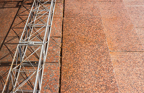 White metal scaffolding casting a shadow on red granite pavement with visible joints and cracks, creating a contrast between industrial and natural materials