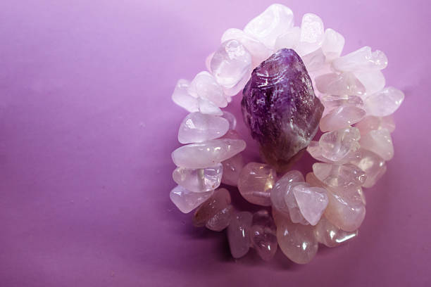 A collection of natural stones, including a polished pink gemstone bracelet and a raw purple amethyst crystal, placed on a lavender-colored fabric backdrop. The soft pink stones are smooth and rounded, while the amethyst has rough, jagged edges with a deep purple hue and lighter inclusions. The contrasting textures and colors create a visually appealing composition.