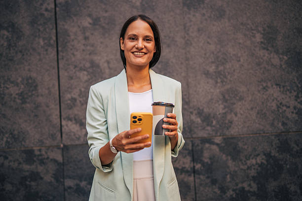 istock Smiling businesswoman using smartphone and drinking coffee against gray wall 2233006150