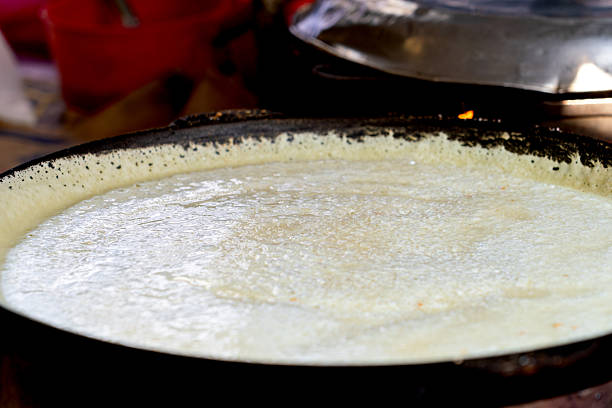 Thin batter spreads evenly on a hot griddle, forming a crepe or dosa in a vibrant kitchen cooking scene.