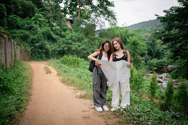 Two friends exploring nature with a map, standing on a dirt trail in a lush green landscape surrounded by trees and hills on a cloudy day