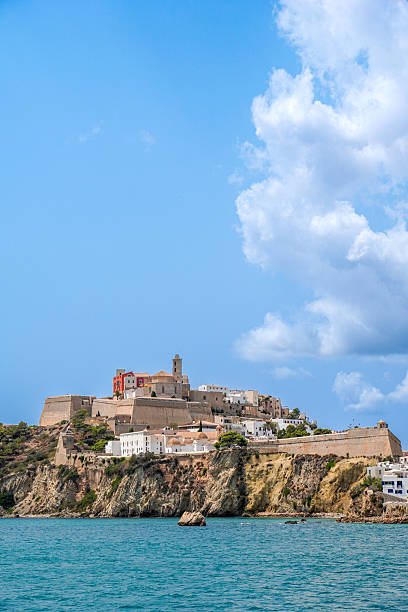 A picturesque view of a historical castle perched on a cliff, surrounded by tranquil waters under a clear blue sky. Eivissa, Ibiza, Spain.