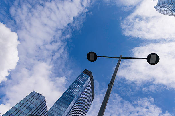 A towering glass skyscraper rises towards the sky, featuring reflections of fluffy white clouds and blue sky on its reflective surface. A lamp post is seen nearby, adding an urban touch to this perspective-enhanced image capturing architectural greatness.
