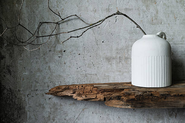 Minimalist still life with a white ceramic vase holding dry branches, placed against a rustic wooden board and textured concrete wall. Natural wabi-sabi style composition.