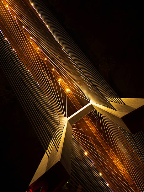 Stunning night view of a cable-stayed bridge lit by warm lights, showcasing modern architectural beauty. The composition emphasizes geometry and nighttime urban design. The glowing lights and intricate cable arrangements give a sense of movement and dynamism.