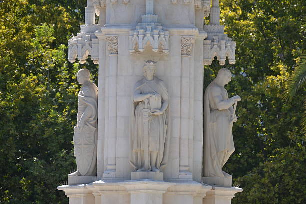 istock A symmetrical close-up of a carved marble structure. Three figures are visible, including a central crowned figure and two others in classical attire. The detailed sculpture is set against a vibrant green foliage background. 2233065997
