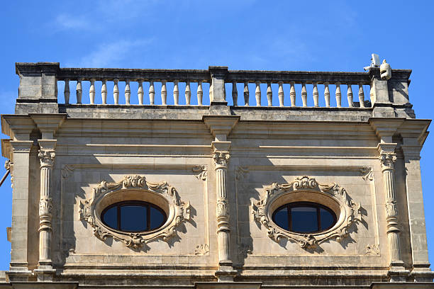 istock A low-angle view of a stone building with a balustrade. The facade features two identical oval windows framed by intricate scrolls and carvings, with fluted columns between them. A clear blue sky is visible above. 2233066579
