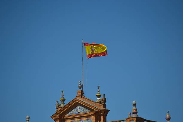 A **Spanish flag** flutters against a clear **blue sky**. The flag is mounted on a pole at the peak of an ornate **brick building**. The roofline is decorated with a tiled pediment and finials.