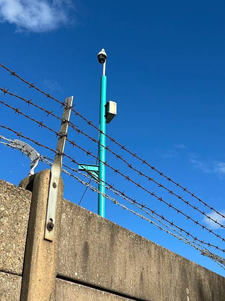 istock Barbed Wire Fence Against Clear Blue Sky with Security Camera in background 2233070809