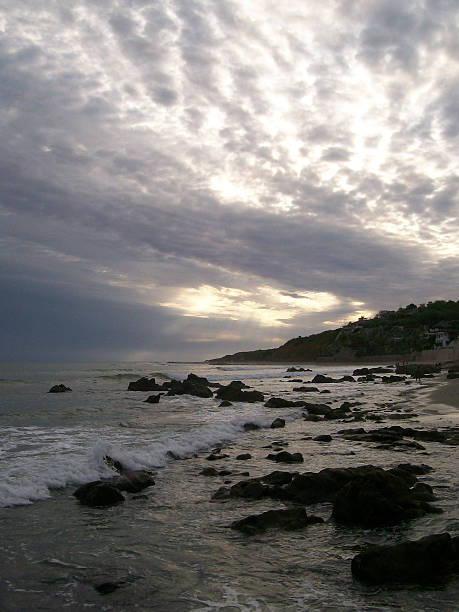 Waves crashing against the rocky shore of General Villamil Playas in Guayas, Ecuador create a stunning scene at sunset, with a dramatic sky filled with clouds enhancing the picturesque view