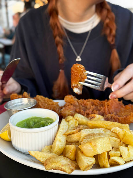 istock Close-up image unrecognisable redheaded woman with braids in restaurant setting eating English fish supper, fish and chips (battered cod) on white plate served with ramekin of mushy peas and small stainless steel bowl of tartar sauce, lemon slice 2233076878