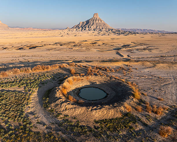 A striking aerial view of Factory Butte rising above the barren Utah desert, with a small reflective pond creating a rare oasis in the arid landscape.