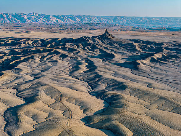 Sweeping aerial view of eroded desert ridges leading toward a distant butte in Utah, with soft evening light accentuating the textures of the barren land.
