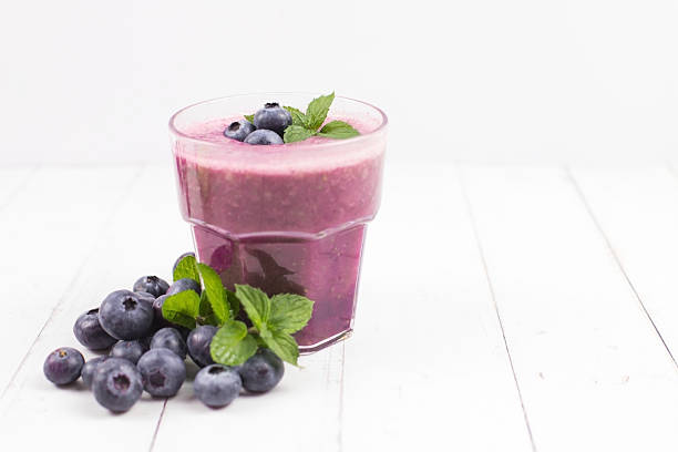 On a light wooden table, a glass of blueberry smoothie is accompanied by fresh blueberries scattered around the base. The bright background highlights the rich purple color of the smoothie and the natural freshness of the berries, perfect for healthy and vibrant beverage photography. High quality photo