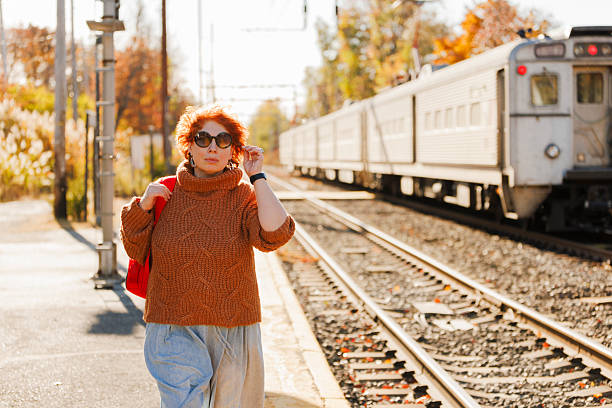 A confident 46-year-old woman with red hair pauses during her afternoon commute, bathed in golden autumn sunlight. Her brown turtleneck sweater complements the fall colors of surrounding trees while she adjusts her sunglasses and holds a red bag. The suburban railway setting features a white commuter train backdrop, with warm backlighting creating an authentic lifestyle moment against the seasonal foliage.
