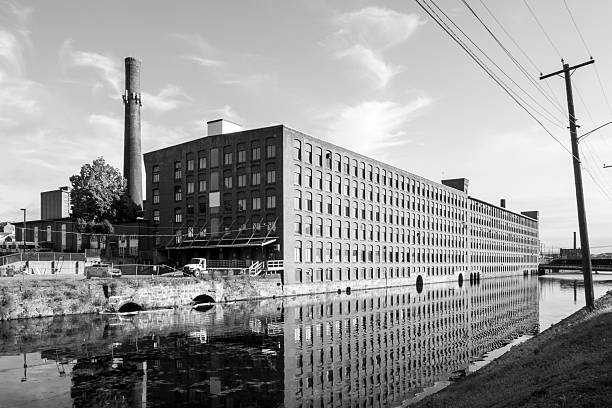 Holyoke, MA - US - Aug 24, 2025 Black and white view of a long brick mill in Holyoke reflects in a calm canal under a bright blue sky, framed by smokestacks and power lines.