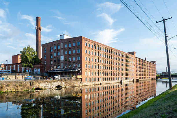 Holyoke, MA - US - Aug 24, 2025 A long brick mill in Holyoke reflects in a calm canal under a bright blue sky, framed by smokestacks and power lines.